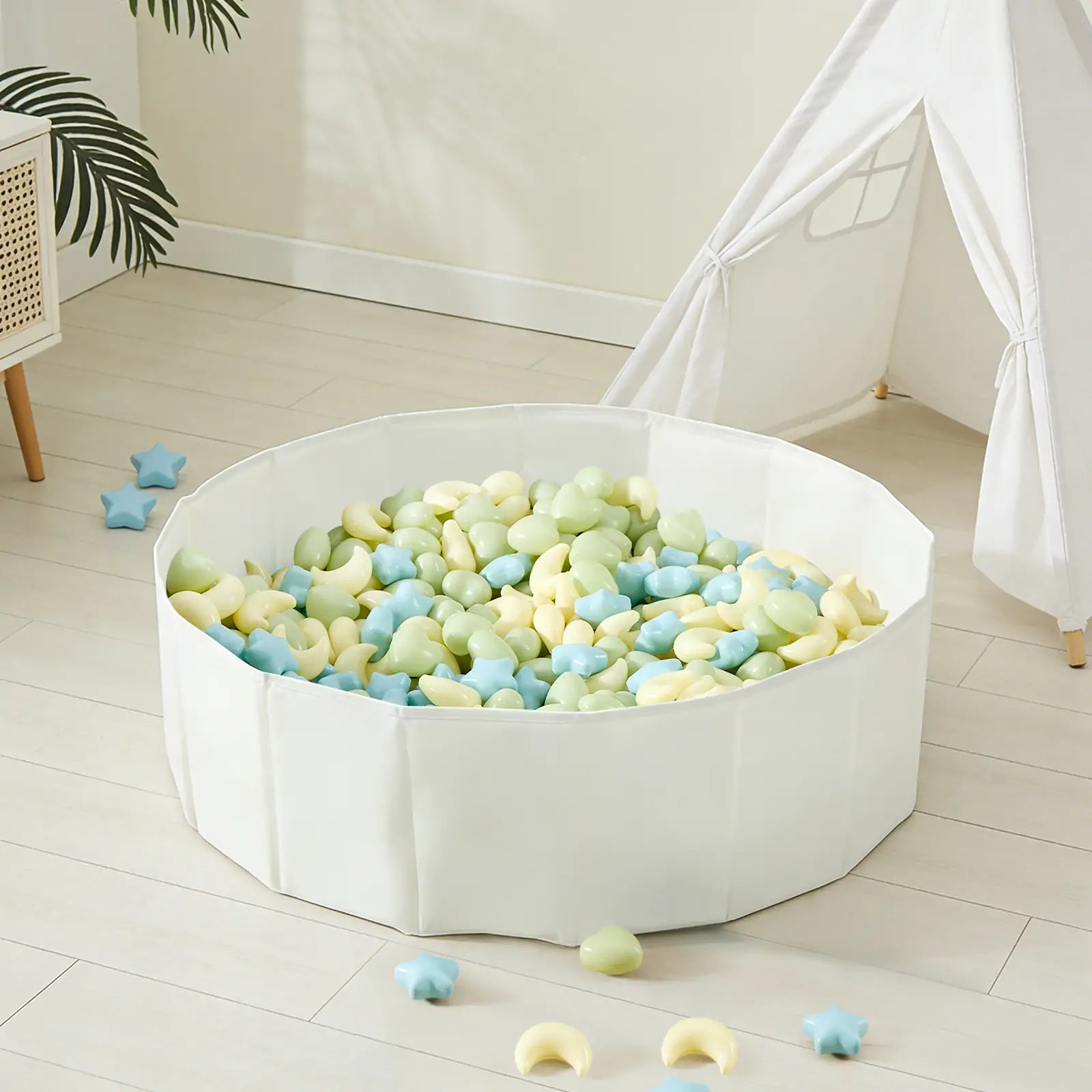 A white playpen filled with Tiny Land® Fun-Shapes Ocean Ball - 200 pcs in pastel green, yellow, and blue and star-shaped toys sits on a light wooden floor near a white teepee tent and a plant. Some Tiny Land balls are scattered outside.|Fun-Shapes
