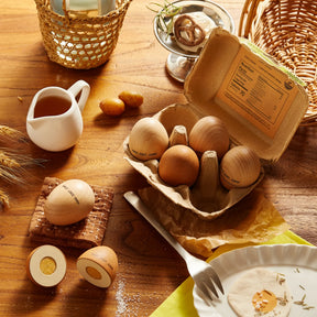The Tiny Land Play Food Egg Combo Playset is displayed on a wooden table with kitchen items like a milk jug, wicker baskets, a plate, a fork, crackers, wheat stalks, and two cut-open wooden eggs with painted yolks.