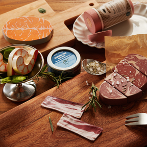A wooden table showcases the Tiny Land® Organic Meat & Seafood Pack by Tiny Land Play Food, featuring realistic steak, bacon, citrus, fish, and a "BLACK PEPPER TUNA" tin—all garnished with rosemary and salt for playful presentation.