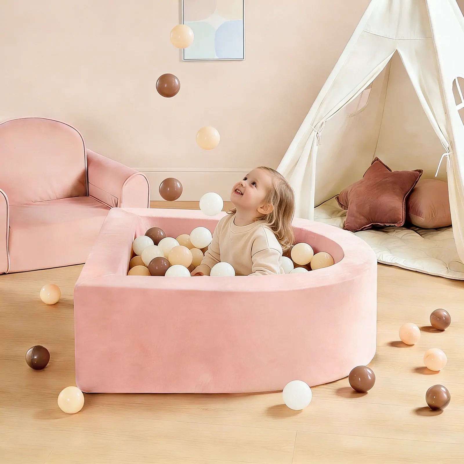 A young child sits in the pink ball pit of the Tiny Land® PlayNest Climbing & Ball Pit Set, smiling and gazing up at several colorful balls tossed into the air. A matching tent, soft climbing blocks, and an armchair are visible in the background of the bright, cozy room, creating a cheerful indoor playground atmosphere by Tiny Land.|Pink