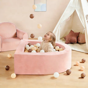 A young child sits in the pink ball pit of the Tiny Land® PlayNest Climbing & Ball Pit Set, smiling and gazing up at several colorful balls tossed into the air. A matching tent, soft climbing blocks, and an armchair are visible in the background of the bright, cozy room, creating a cheerful indoor playground atmosphere by Tiny Land.|Pink