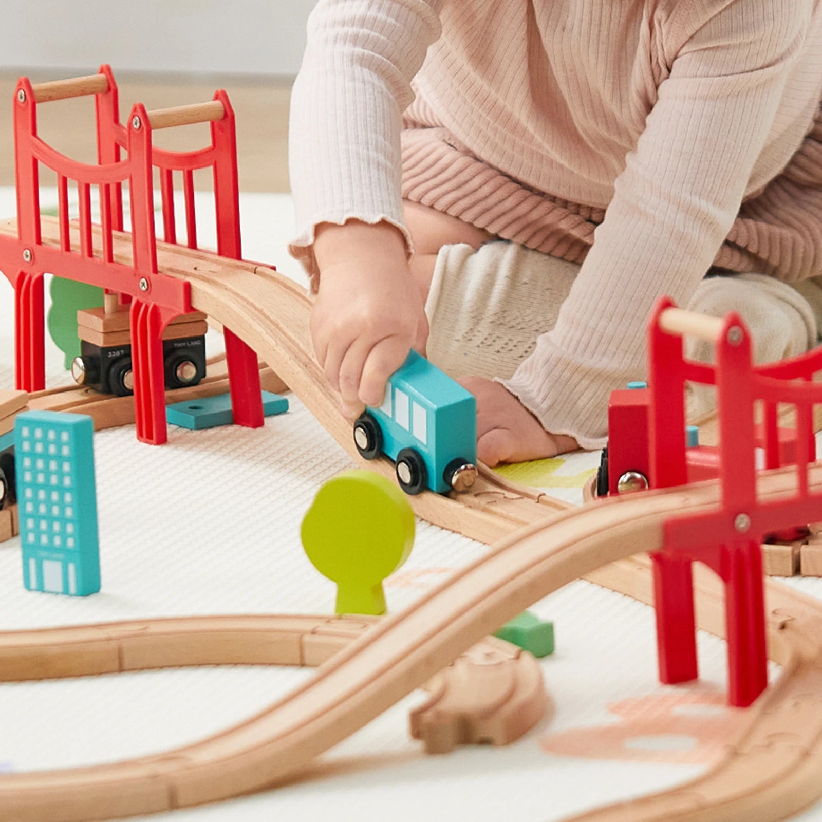 A child in a pink sweater and tights plays with the Tiny Land® Wooden Train Set for Children 39 Pcs, guiding a blue train over tracks and bridges, surrounded by toy buildings and trees.|39 Pcs