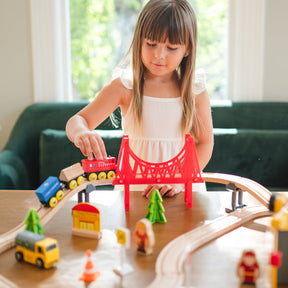 A young girl plays with the Tiny Land® Wooden Train Set for Children (39 pcs) on a table. The eco-friendly set by Tiny Land includes a red bridge, trees, figures, and vehicles. Sunlight streams through the windows behind her.|39 Pcs