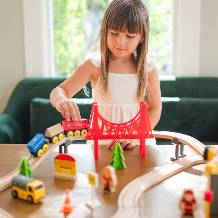 A young girl plays with the Tiny Land® Wooden Train Set for Children (39 pcs) on a table. The eco-friendly set by Tiny Land includes a red bridge, trees, figures, and vehicles. Sunlight streams through the windows behind her.|39 Pcs