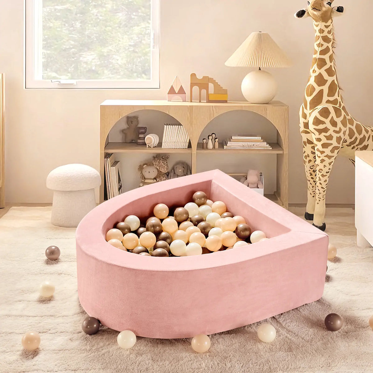 A cozy, pastel toddler play space features the Tiny Land® PlayNest Ball Pit with 200 ocean balls on a cream rug. A toy giraffe stands by a bookshelf with books and decor as soft, natural light streams in from the window.
