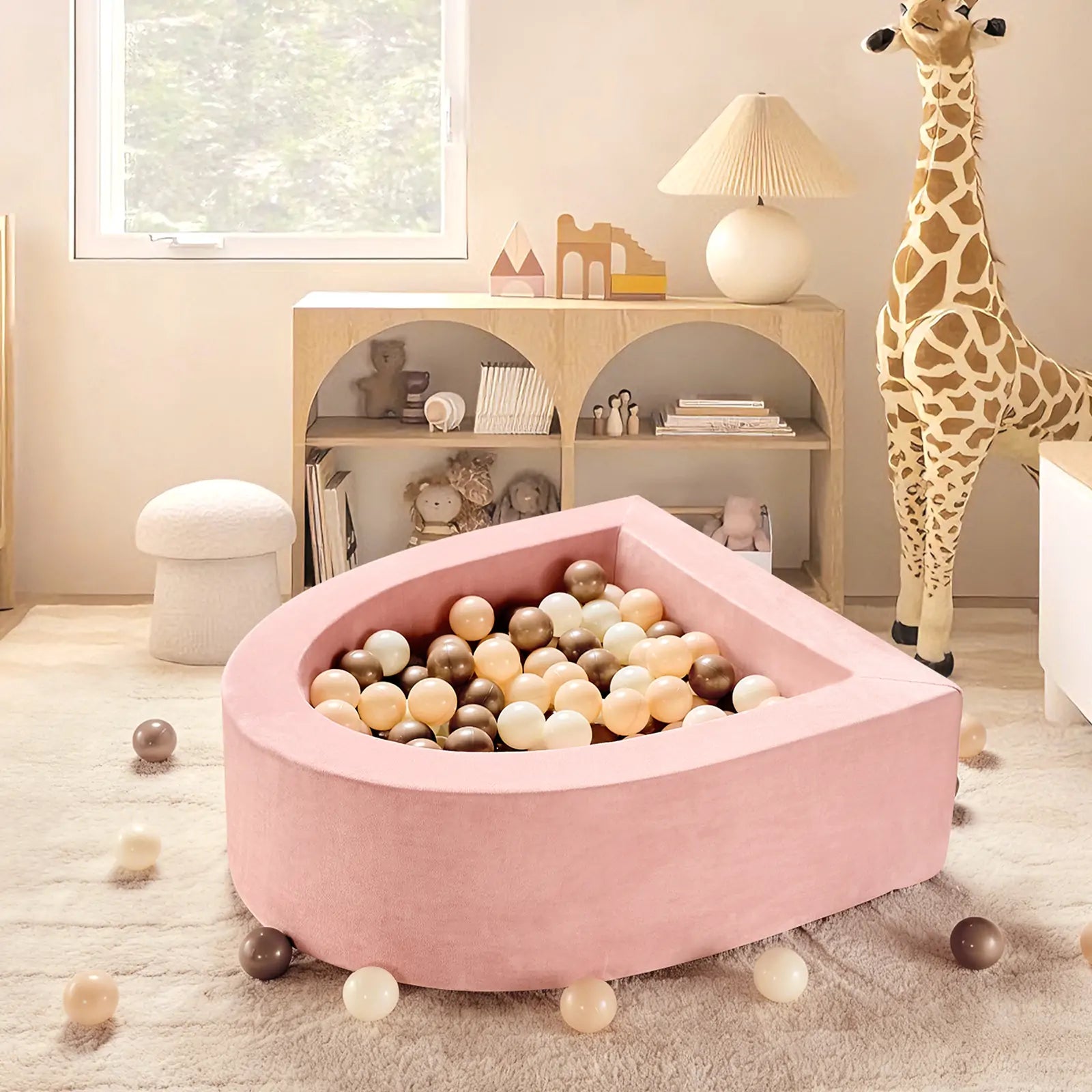 A cozy, pastel toddler play space features the Tiny Land® PlayNest Ball Pit with 200 ocean balls on a cream rug. A toy giraffe stands by a bookshelf with books and decor as soft, natural light streams in from the window.