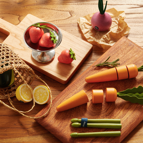 A Tiny Land Play Food Organic Fruit & Veggie Pack sits on the table, with toy strawberries in a glass, a lemon slice, carrot pieces on a board, green tongs, and a purple veggie—realistic play food for toddlers artfully arranged.