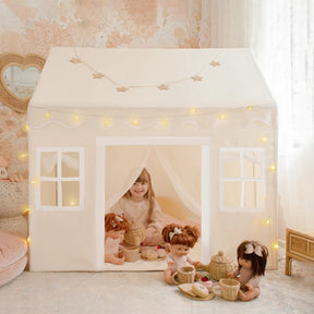 A young child sits inside the Tiny Land® Playhouse Tent in natural cream, surrounded by dolls and toy picnic baskets. Fairy lights, star garlands, and sheer curtains create a cozy, whimsical scene with this magical Tiny Land indoor tent|white