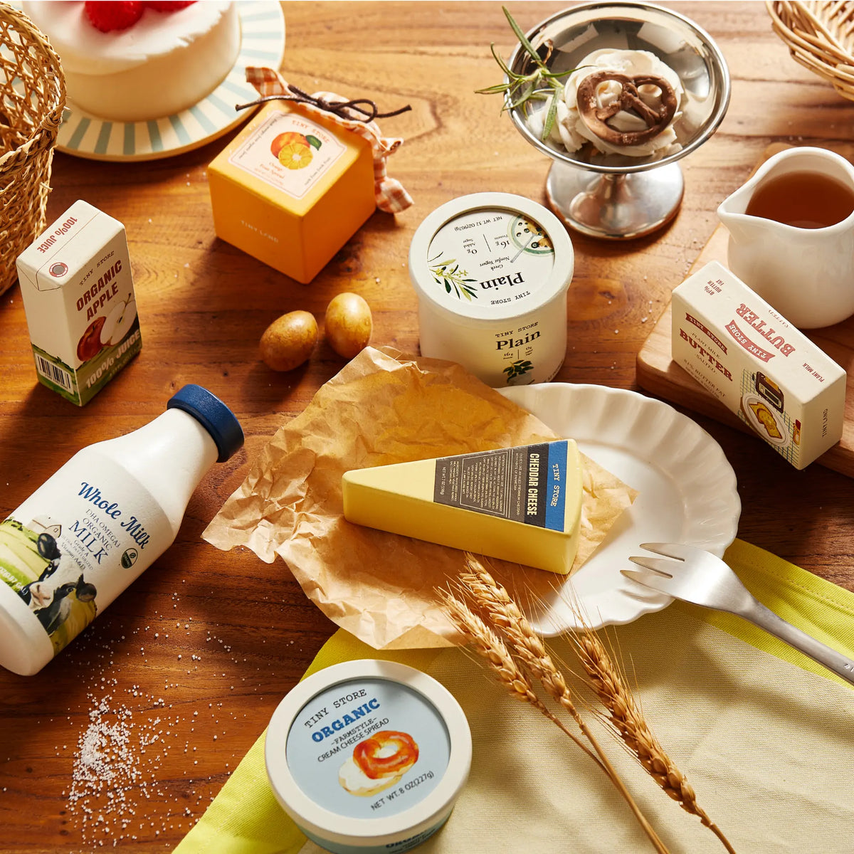 A wooden table displays Tiny Land Play Food’s Tiny Land® Organic Dairy Pack, featuring a wedge of cheese on parchment, milk, yogurt, butter, eggs, and packaged goods for realistic play food fun for toddlers. Wheat stalks are nearby.