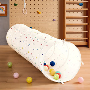 The Tiny Land® Adventure Play Tunnel - Colorful Polka Dots from Tiny Land sits on a wooden floor, filled with multicolored balls. In front of a climbing wall, gym rings, ladder, and children’s art, it creates a vibrant indoor play space.