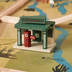 A wooden Texaco gas station toy with a red fuel pump rests on the Tiny Land® 1920 Railway Adventure Train Table play map, featuring roads and rivers. Wooden train tracks and a blurred toy figure appear in the background. Brand: Tiny Land.