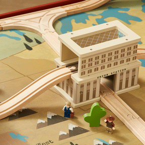 The Tiny Land® 1920 Railway Adventure Train Table features colorful, stylized train tracks winding through a Union Station city model on a map-themed play mat, plus two toy figures and a green tree for interactive fun.