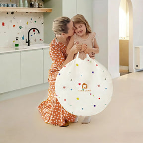 A woman in a polka dot dress kneels and hugs a smiling young girl, who holds a round white bag with colorful dots and the text “TINY LAND,” in a modern kitchen—the perfect setting for fun activities with the Tiny Land® Adventure Play Tunnel by Tiny Land.|Colorful Dots