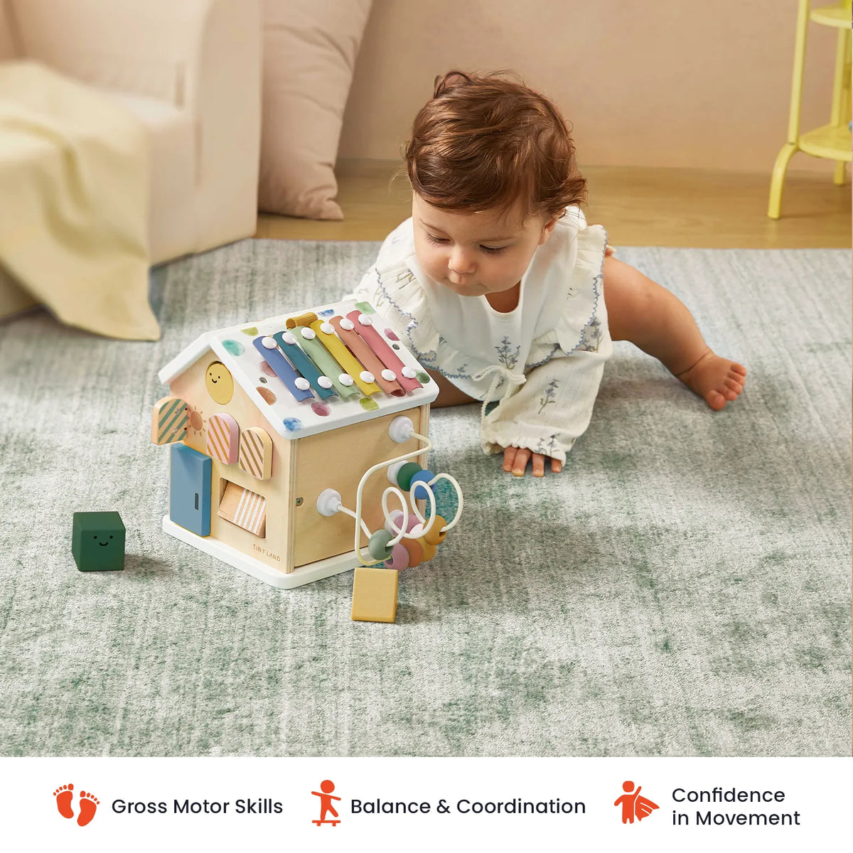 A baby explores the Complete Development Playset — Baby Walker + Activity Cube by Tiny Land, engaging on a soft rug with this colorful wooden Montessori-inspired activity house. Icons and text below highlight how it supports gross motor skills, fine motor skills, balance and coordination, and confidence in movement.