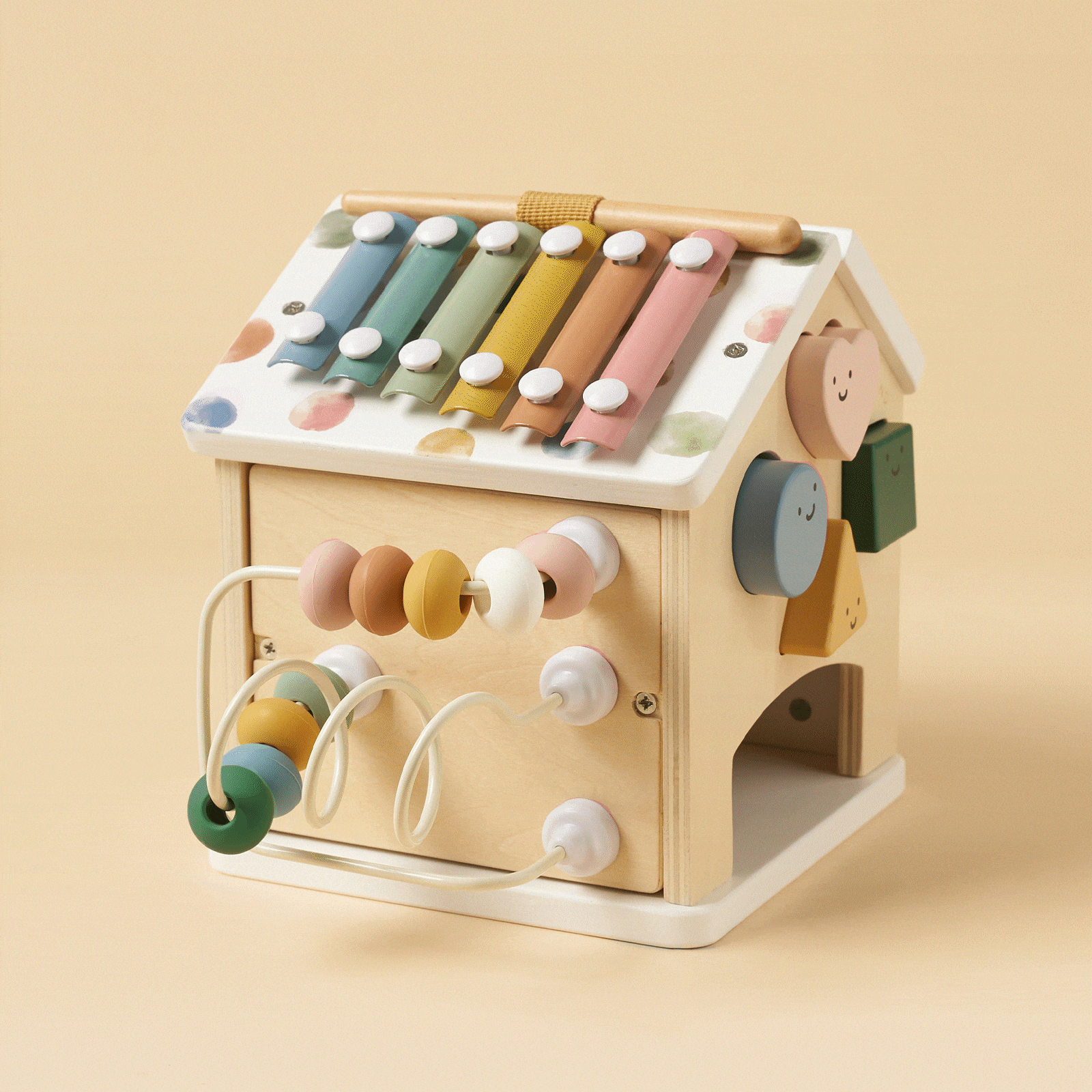 The Tiny Land Cozy Learning Playset combines a playmat with a wooden house-shaped Montessori activity cube featuring a colorful xylophone roof, bead maze, spinning face blocks, and pastel colors on a light beige background.