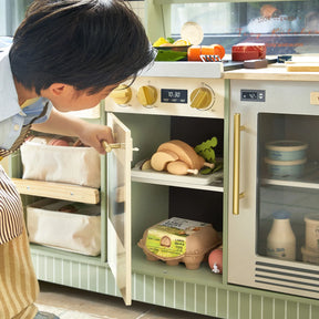 A child in an apron opens the Tiny Land® BistroSwitch Play Kitchen, revealing toy food like wooden bread and eggs. This realistic set by Tiny Land features detailed knobs, display screens, and utensils for imaginative pretend play.|Green