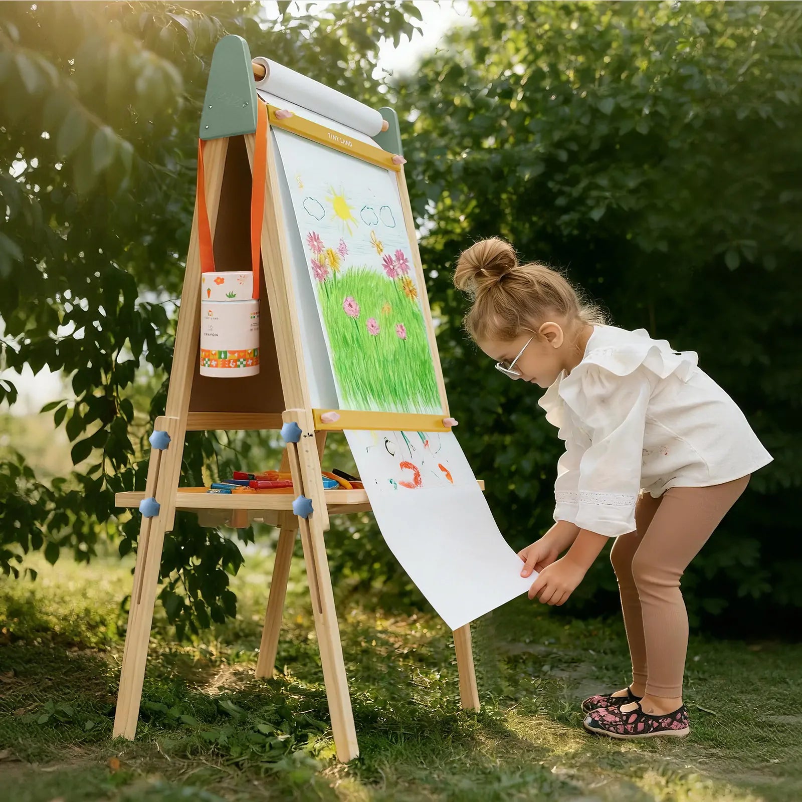 A young girl with glasses creates art featuring flowers, sunshine, and butterflies at the Tiny Land® 3-in-1 Art Easel for Kids - Morandi by Tiny Land, outdoors on grass surrounded by green bushes and trees.|green