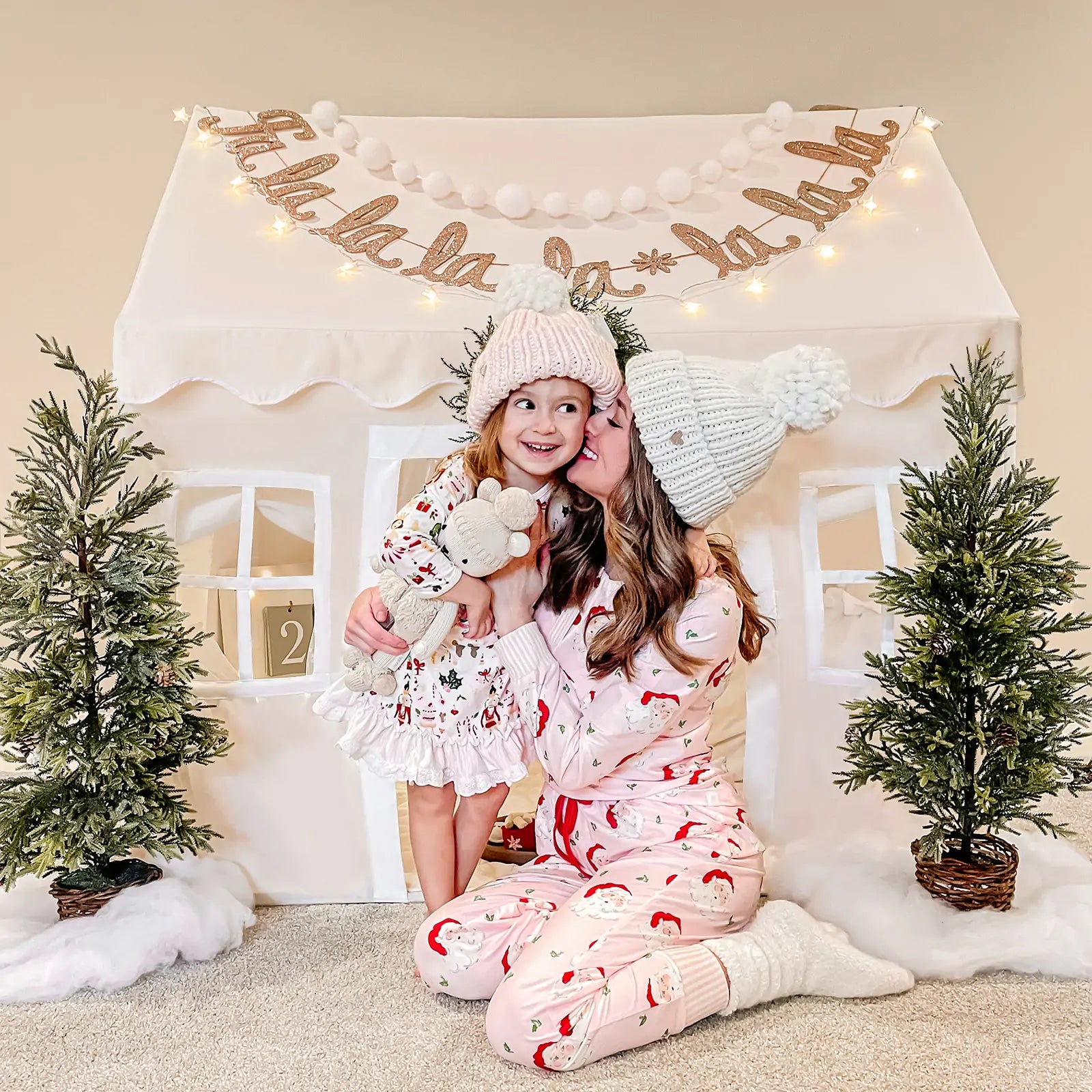 A woman and young girl in festive pajamas smile and embrace by a Tiny Land Bright Play & Art Set—Colorful Easel + Cream Playhouse with Lights—decorated with holiday trees, as the girl hugs a stuffed animal|white