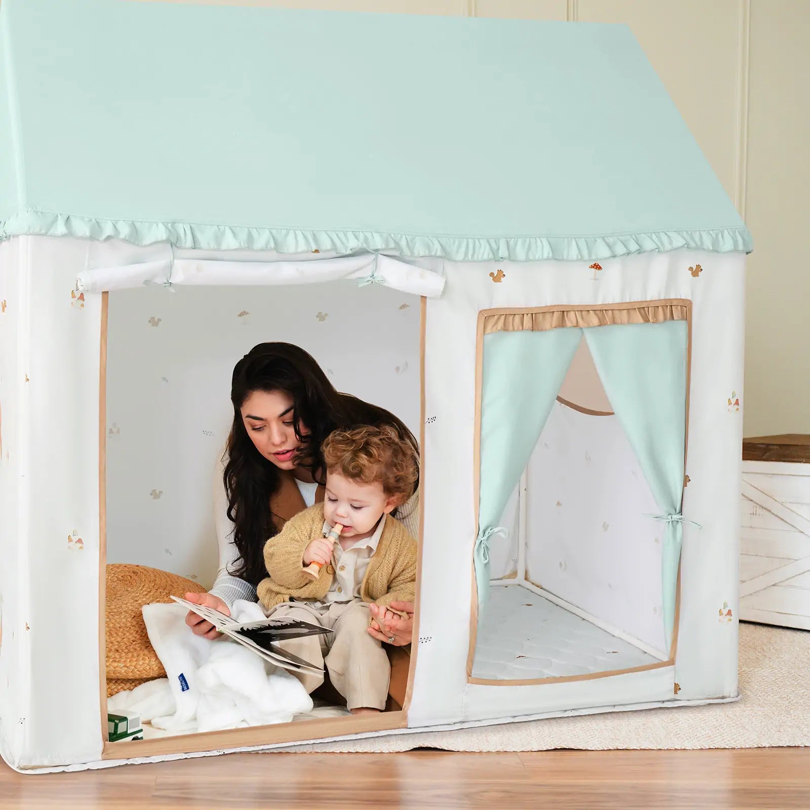 A woman and child sit in the Tiny Land Nature-inspired Play & Art Set — Sage Easel + Forest Play House, shaped like a small house with a mint green roof, reading together. A stuffed toy and blanket rest nearby on the floor.|green