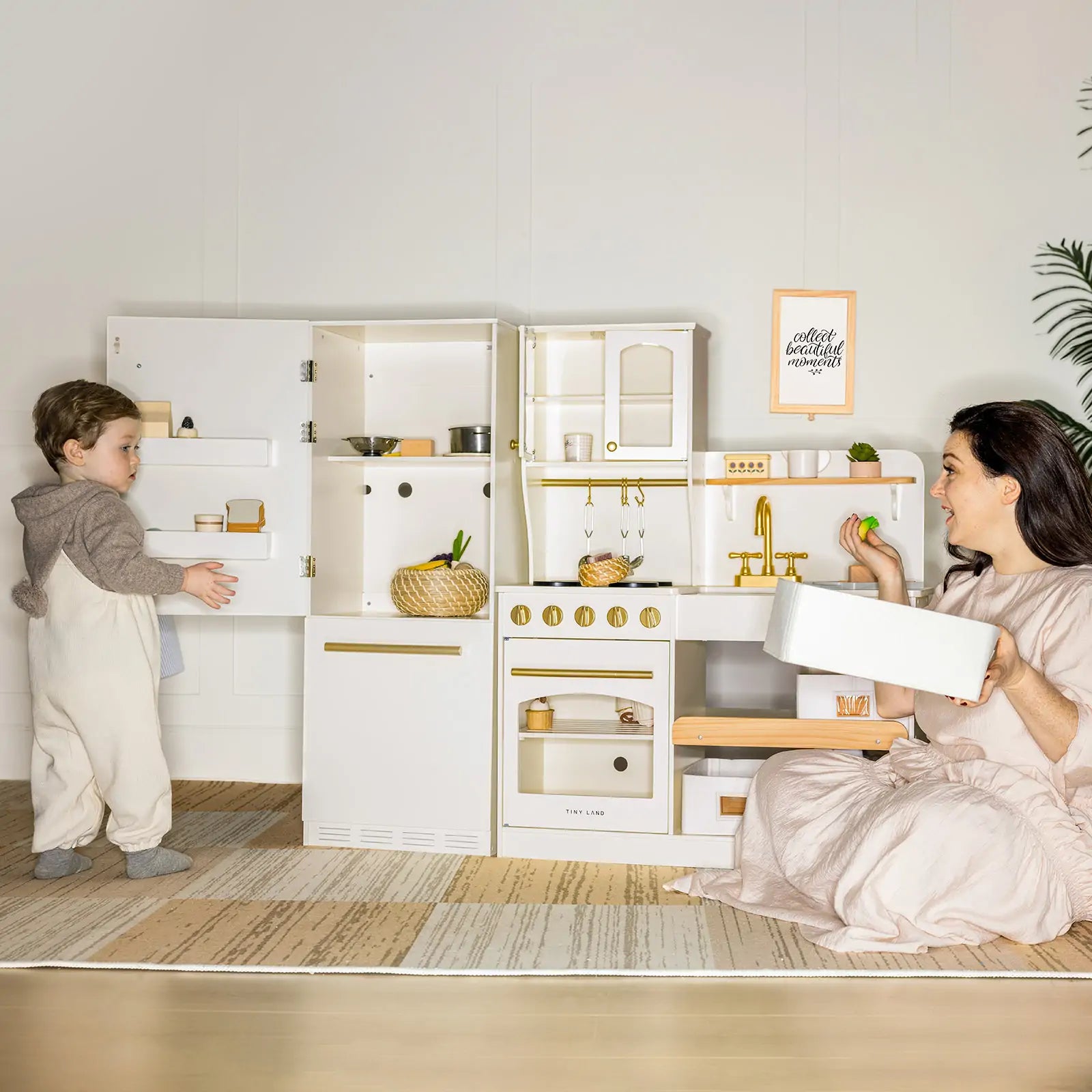 A woman and child enjoy the Tiny Land® Play Kitchen Accessories – Wooden Cabinet Refrigerator for Kids. The woman kneels, smiling with a tray, as the child stands nearby in a bright room with minimal decor and a rug.