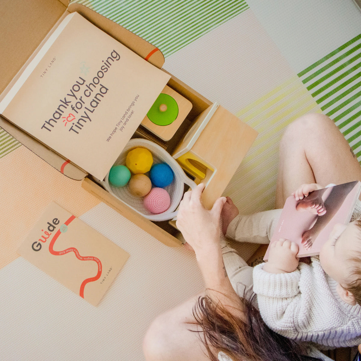 A woman and a baby sit on a play mat with an open Tiny Land® Montessori Toys for Newborns (7–9 Months) box, colorful toys, a thank you card, and guide booklet. The baby smiles while holding a sensory ball from Tiny Land.