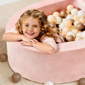A smiling young child with wavy blonde hair leans on the edge of the Tiny Land® PlayNest Ball Pit with 200 Ocean Balls, a cozy toddler play space by Tiny Land, with some pastel balls scattered on the carpet.