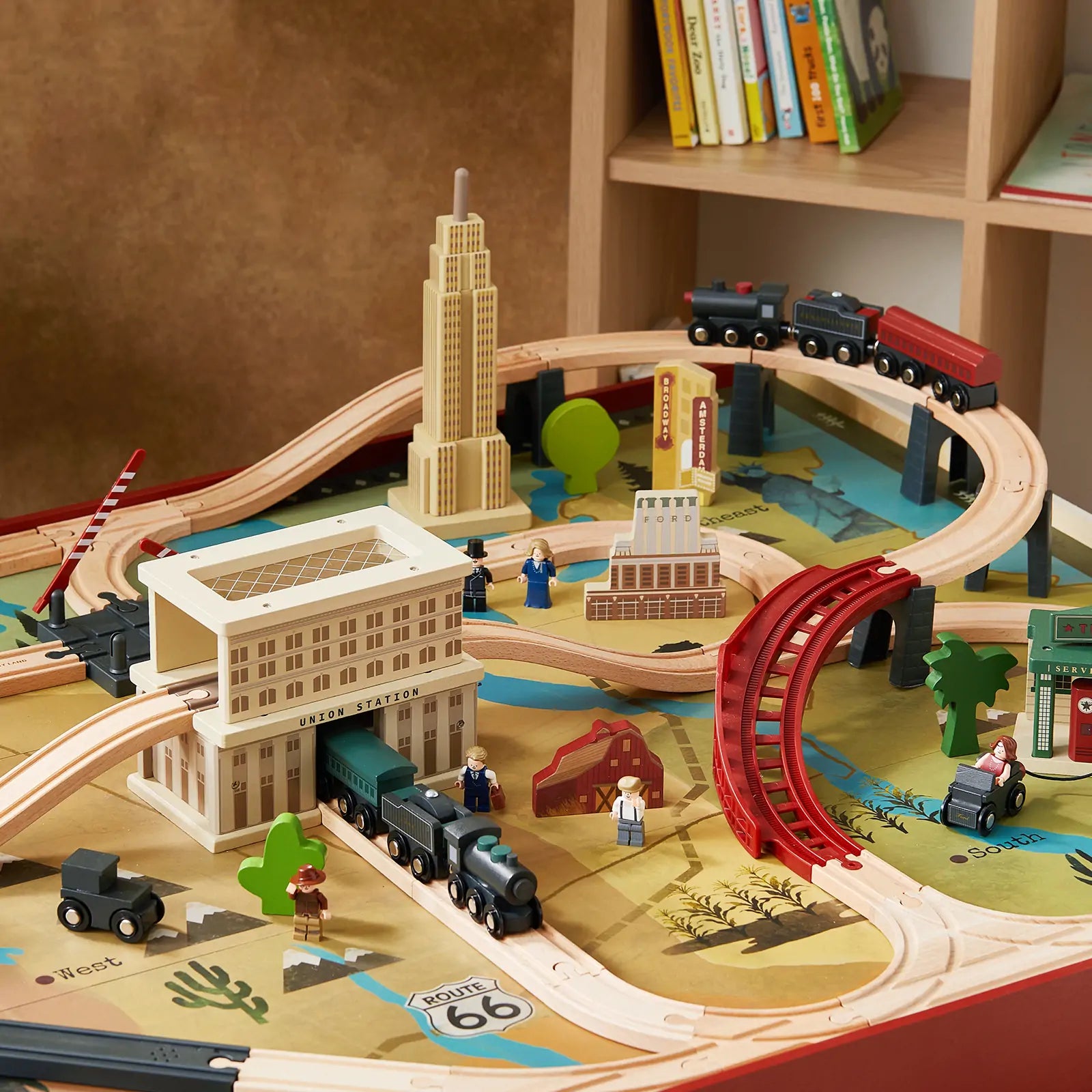 The Tiny Land® 1920 Railway Adventure Train Table by Target features tracks, bridges, buildings, cars, trees, and figurines on a colorful 1920s city play mat. Books can be seen on a shelf in the background.
