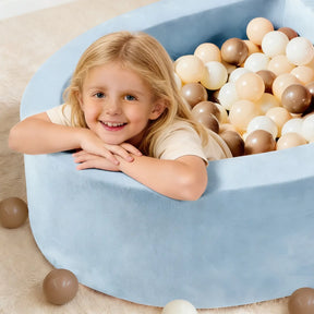 A smiling blonde child leans over the edge of a Tiny Land® PlayNest Ball Pit with 200 ocean balls in beige, white, and brown by Tiny Land, with some balls scattered on a beige carpet outside the high-density foam pit.