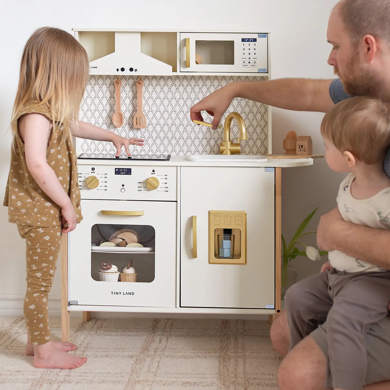 A man and two children enjoy pretend play with the Tiny Land® PopJoy Kitchen & Fridge Set on a beige carpet. The girl stands by the oven, while the man holds the younger child and reaches toward the sink indoors.