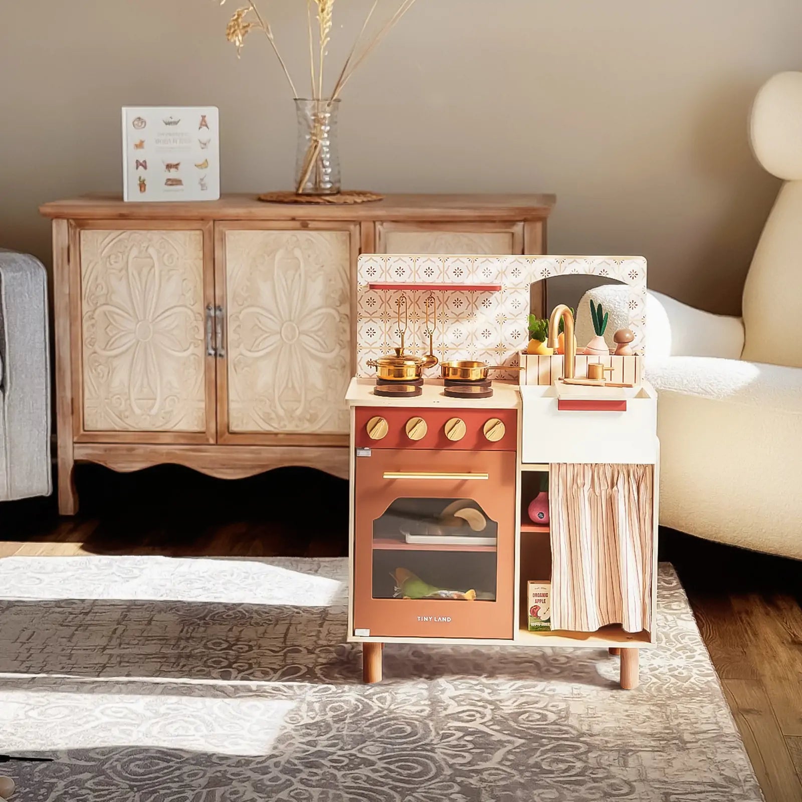 The Tiny Land® Mediterranean Play Kitchen by Tiny Land, complete with toy cookware and vegetables, stands on a patterned rug in a cozy, sunlit living room featuring a wooden cabinet, armchair, and decorative vase in the background|Mediterranean