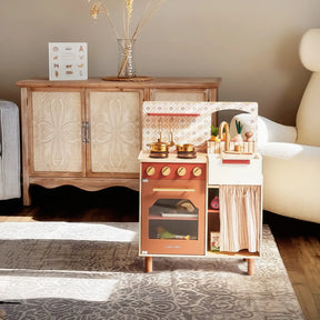 The Tiny Land® Mediterranean Play Kitchen by Tiny Land, complete with toy cookware and vegetables, stands on a patterned rug in a cozy, sunlit living room featuring a wooden cabinet, armchair, and decorative vase in the background|Mediterranean