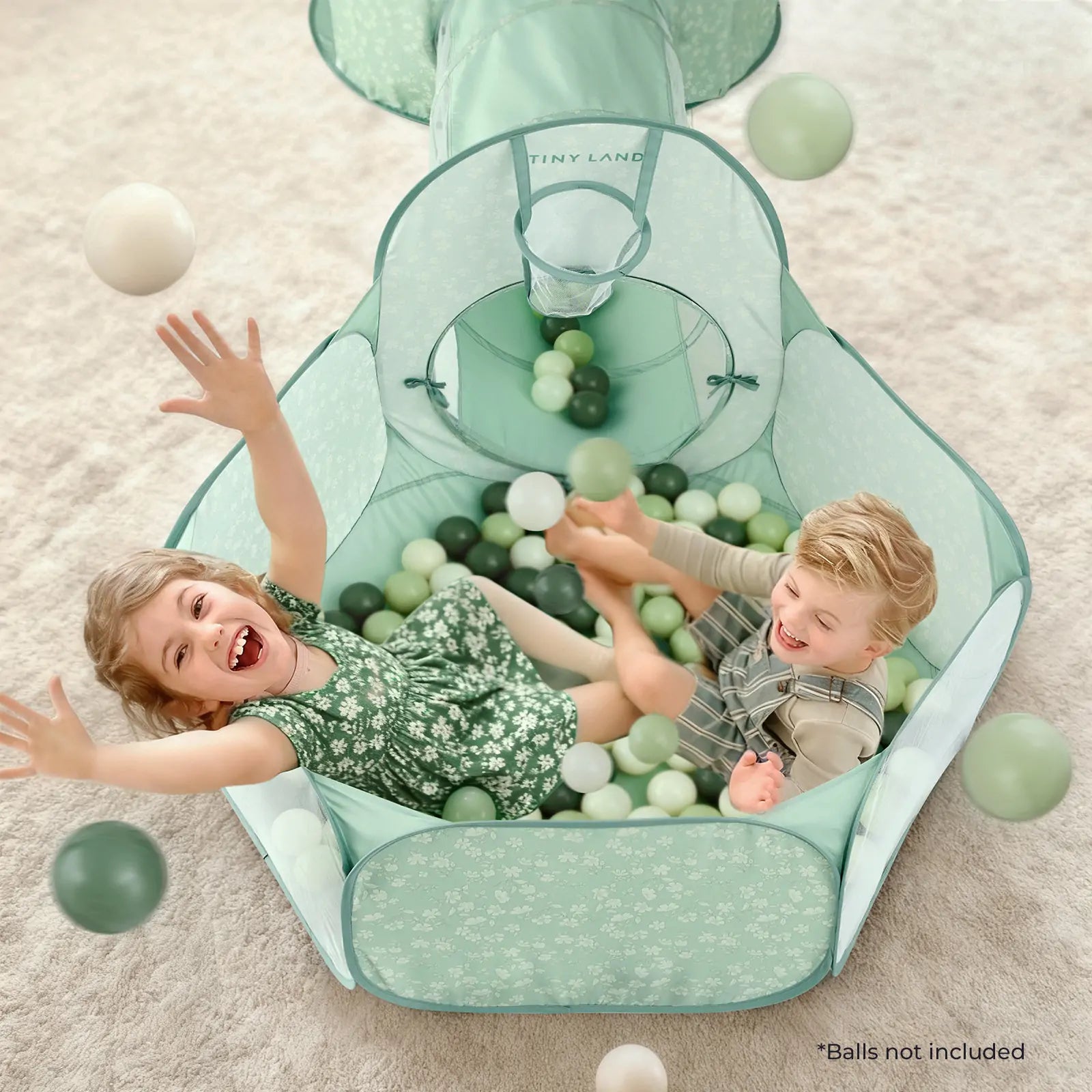 Two young children play and laugh in the mint green Tiny Land® Compact Adventure 3-in-1 Play Tunnel, tossing white and green balls. The ball pit features soft pastel colors and a floral pattern. Text reads: "*Balls not included.|green
