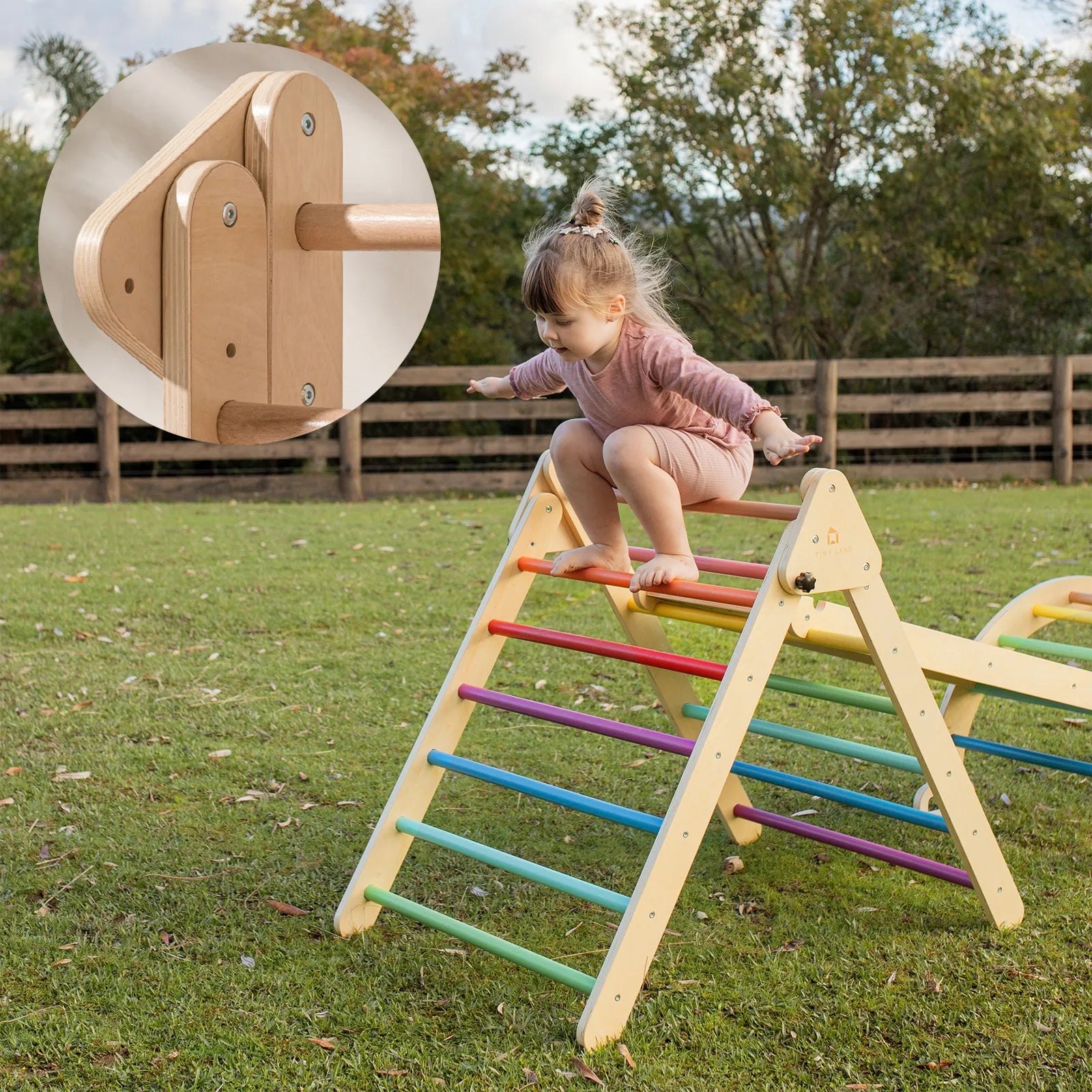 A young child in pink climbs the Tiny Land® Pikler Triangle 7-in-1 Montessori Climbing Set by Tiny Land outdoors on grass. An inset displays sturdy wooden hardware and joints, with trees and a wooden fence visible in the background.