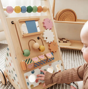 A baby plays with the Tiny Land® Versatile Natural Wooden Baby Walker by Tiny Land, featuring beads, gears, a mirror, and pastel-colored blocks, on a patterned rug in a bright, cozy room.