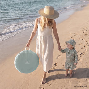A woman in a white dress and sunhat walks on the beach, holding hands with a small child. Nearby, their Tiny Land® Baby Beach Tent & Sand Toy Set from Tiny Land sits ready for play on the sand, with the ocean visible in the background.|Green