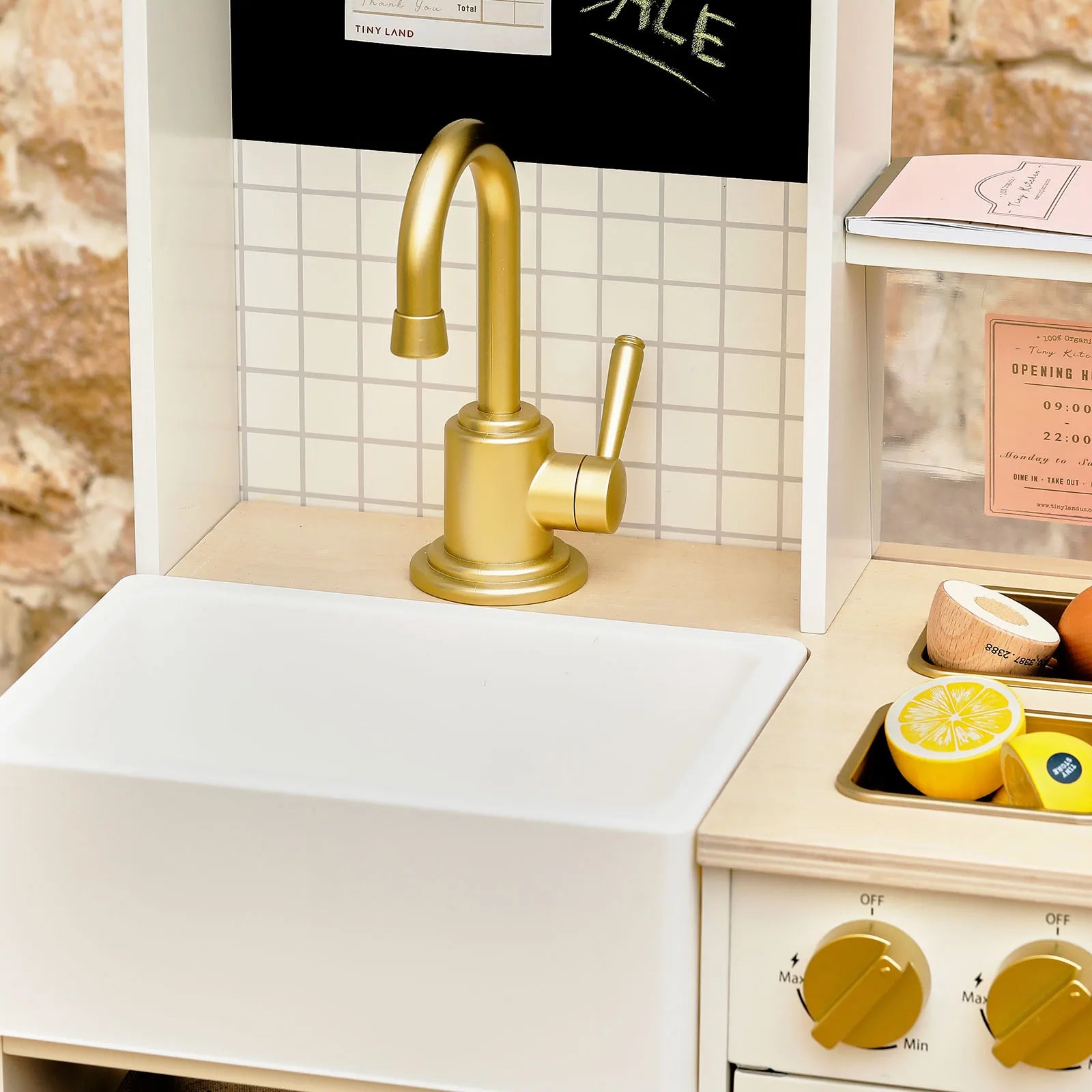 Close-up of the Tiny Land® BistroSwitch Play Kitchen by Tiny Land, featuring a gold-colored faucet, white basin, grid-patterned backsplash, wooden countertop, and sliced toy lemons and fruit near knobs and a pretend oven.|White