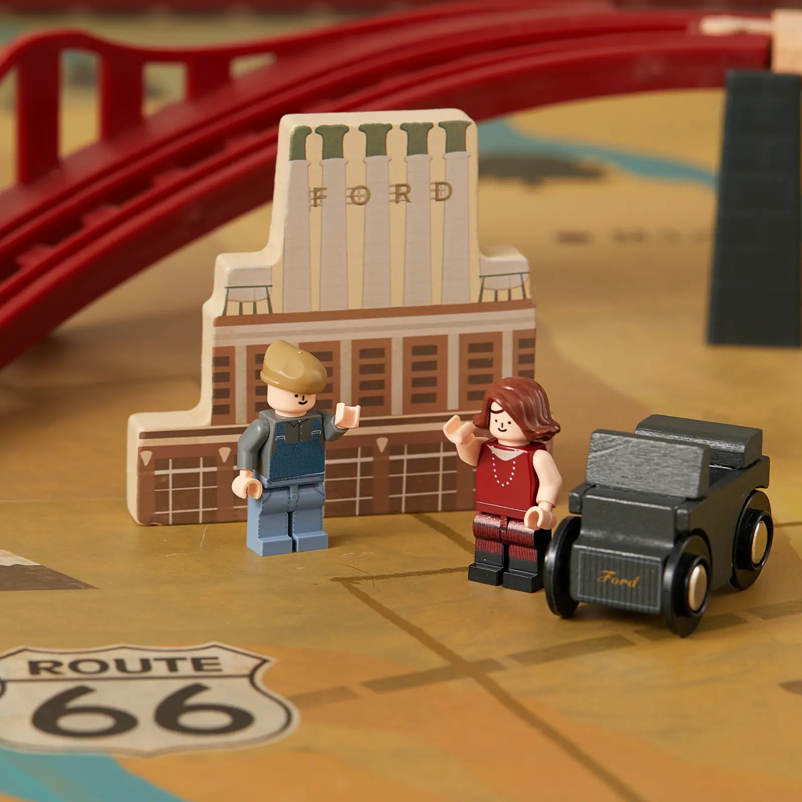 Two LEGO-style figures stand by a vintage black Ford car in front of a wooden building labeled Ford. Behind them, the Tiny Land® 1920 Railway Adventure Train Table from Target features a red toy bridge and Route 66 marking, creating a playful city scene.