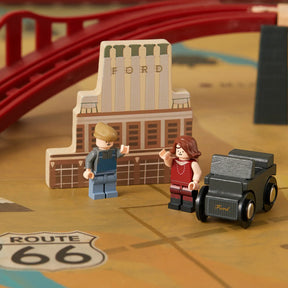 Two LEGO-style figures stand by a vintage black Ford car in front of a wooden building labeled Ford. Behind them, the Tiny Land® 1920 Railway Adventure Train Table from Target features a red toy bridge and Route 66 marking, creating a playful city scene.