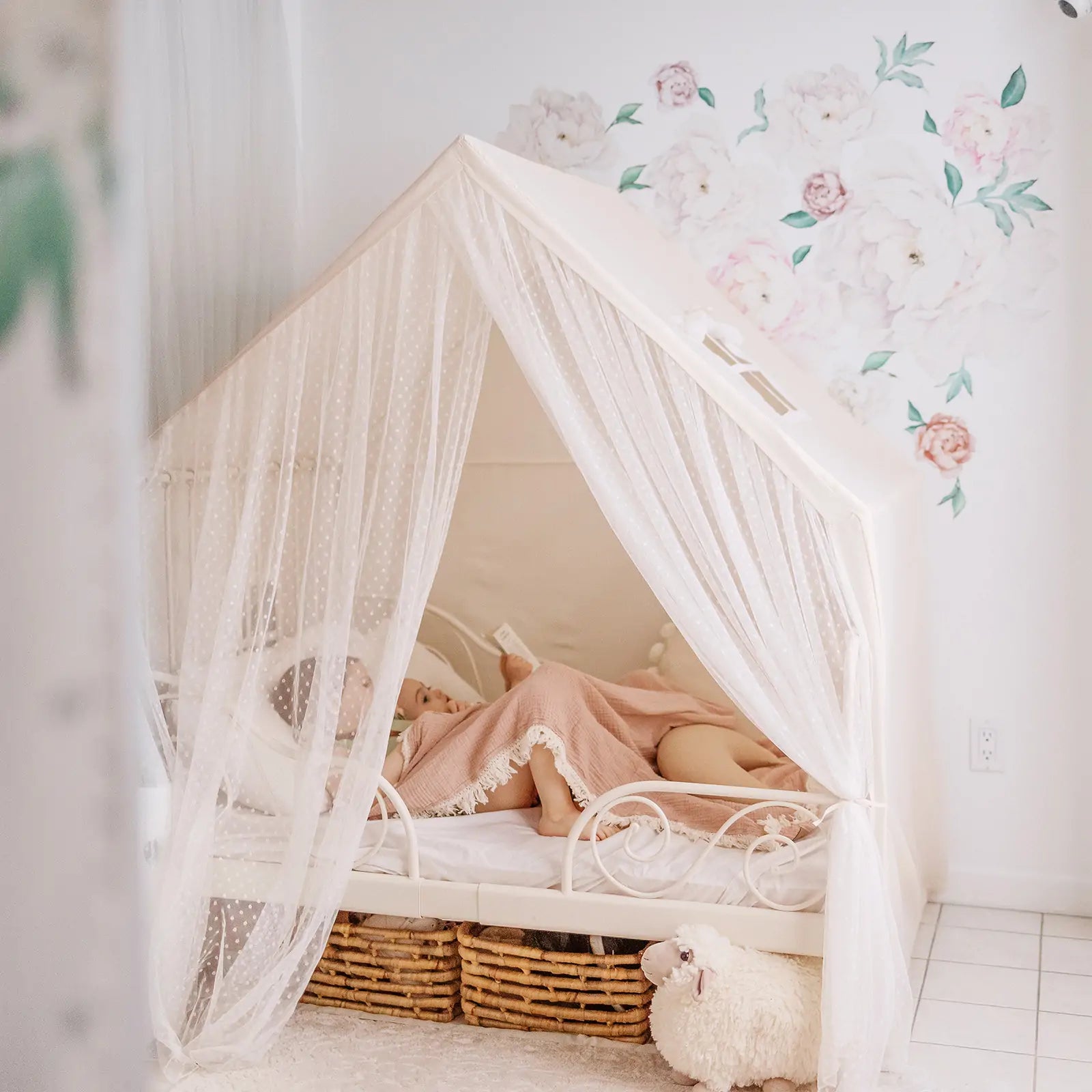 A child rests in the Tiny Land® Chef’s Corner Playset—a cream, house-shaped playhouse with sheer curtains. Floral wall decals, wicker baskets under the bed, and a plush sheep toy on white tiles create a serene atmosphere.|mesh