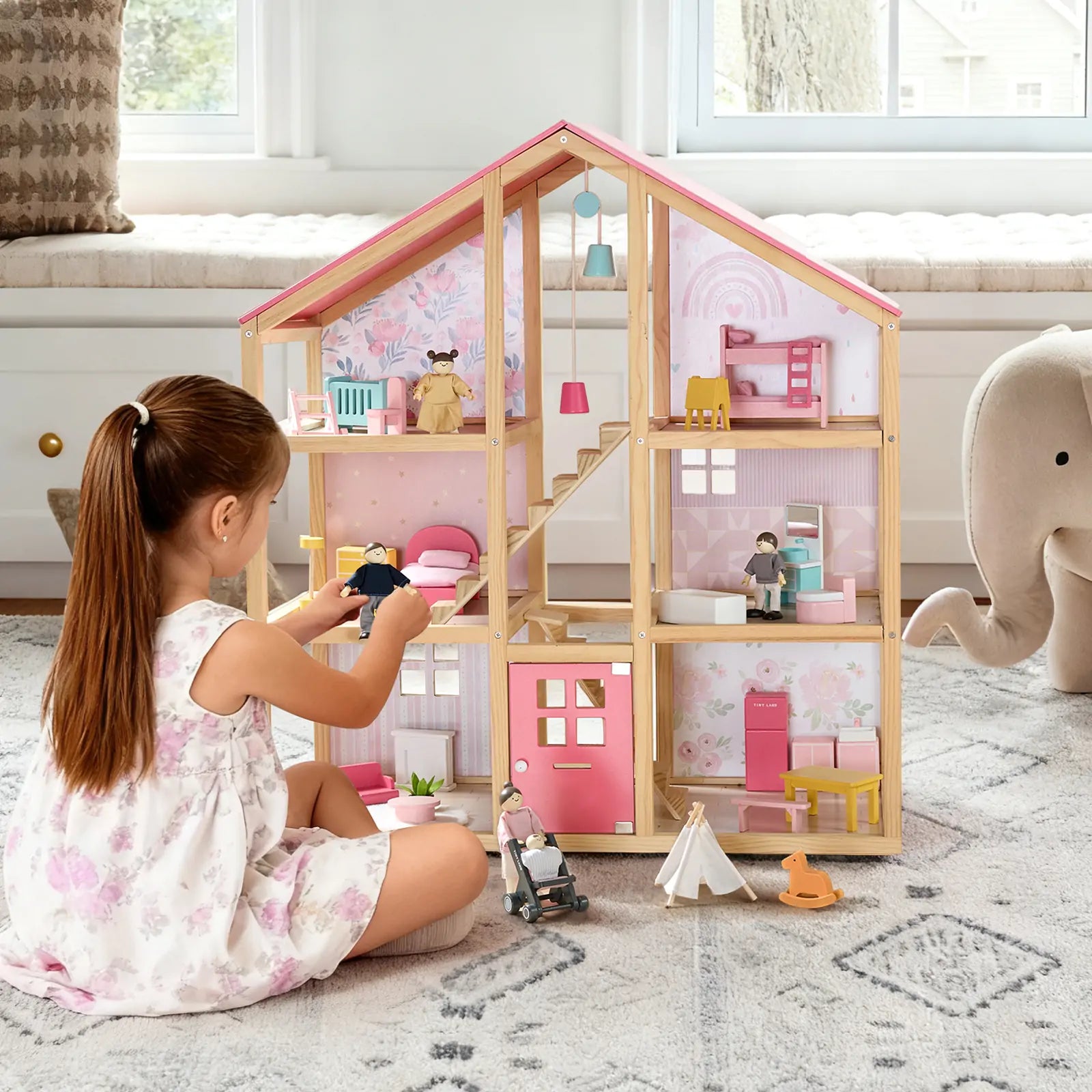 A young girl plays with the Tiny Land® Sweetwood Love Dollhouse & Wooden Dollhouse Dolls, arranging furniture on a patterned rug beside a stuffed elephant in a bright, cozy room.