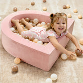A smiling young girl enjoys the Tiny Land® PlayNest Ball Pit with 200 Ocean Balls by Tiny Land, lying in the pink crescent-shaped pit filled with beige, brown, and white balls on a light carpeted playroom floor.