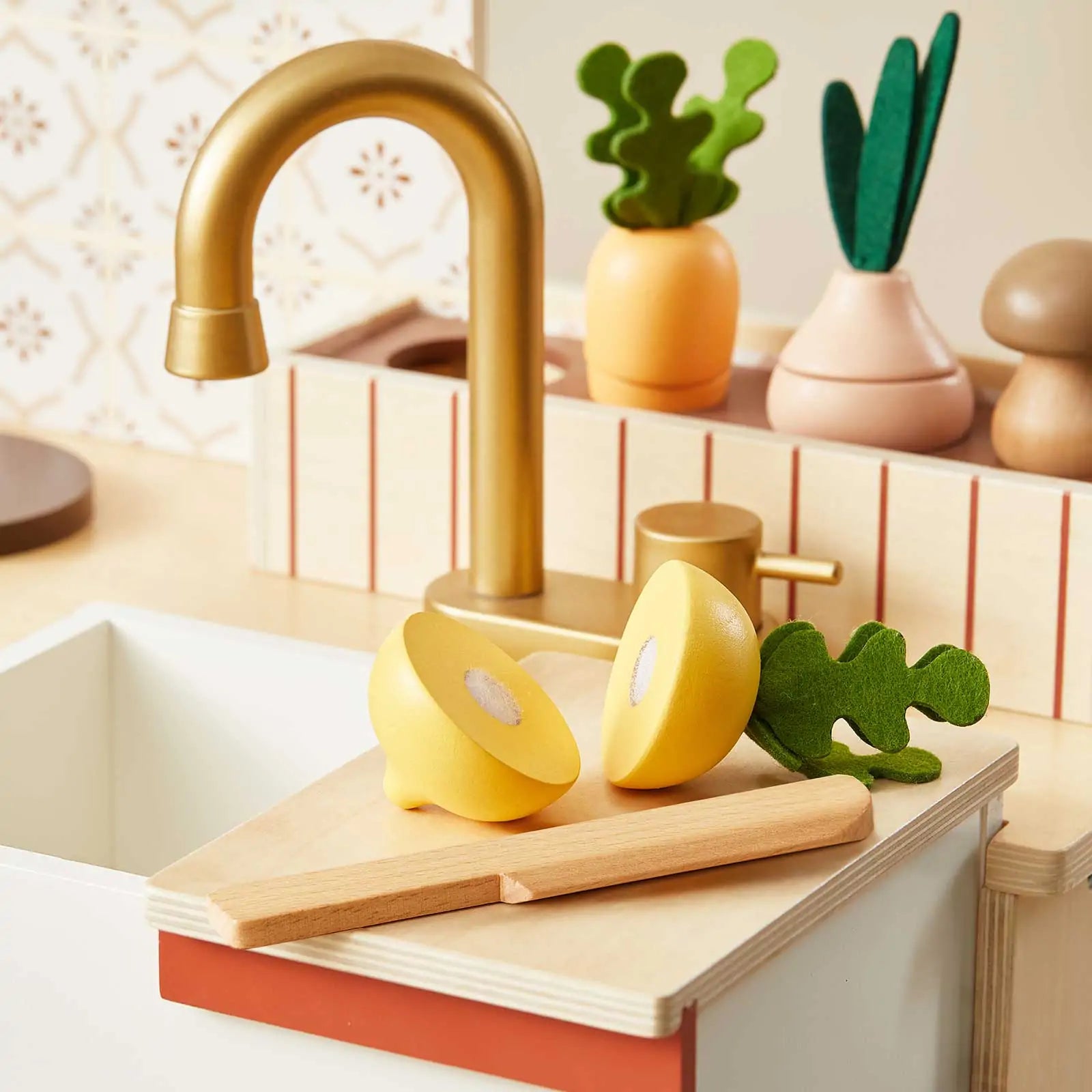 The Tiny Land® Mediterranean Play Kitchen by Tiny Land features a gold faucet, wooden cutting board with knife, two yellow toy fruit halves, and decorative plants in vases on the countertop|Mediterranean