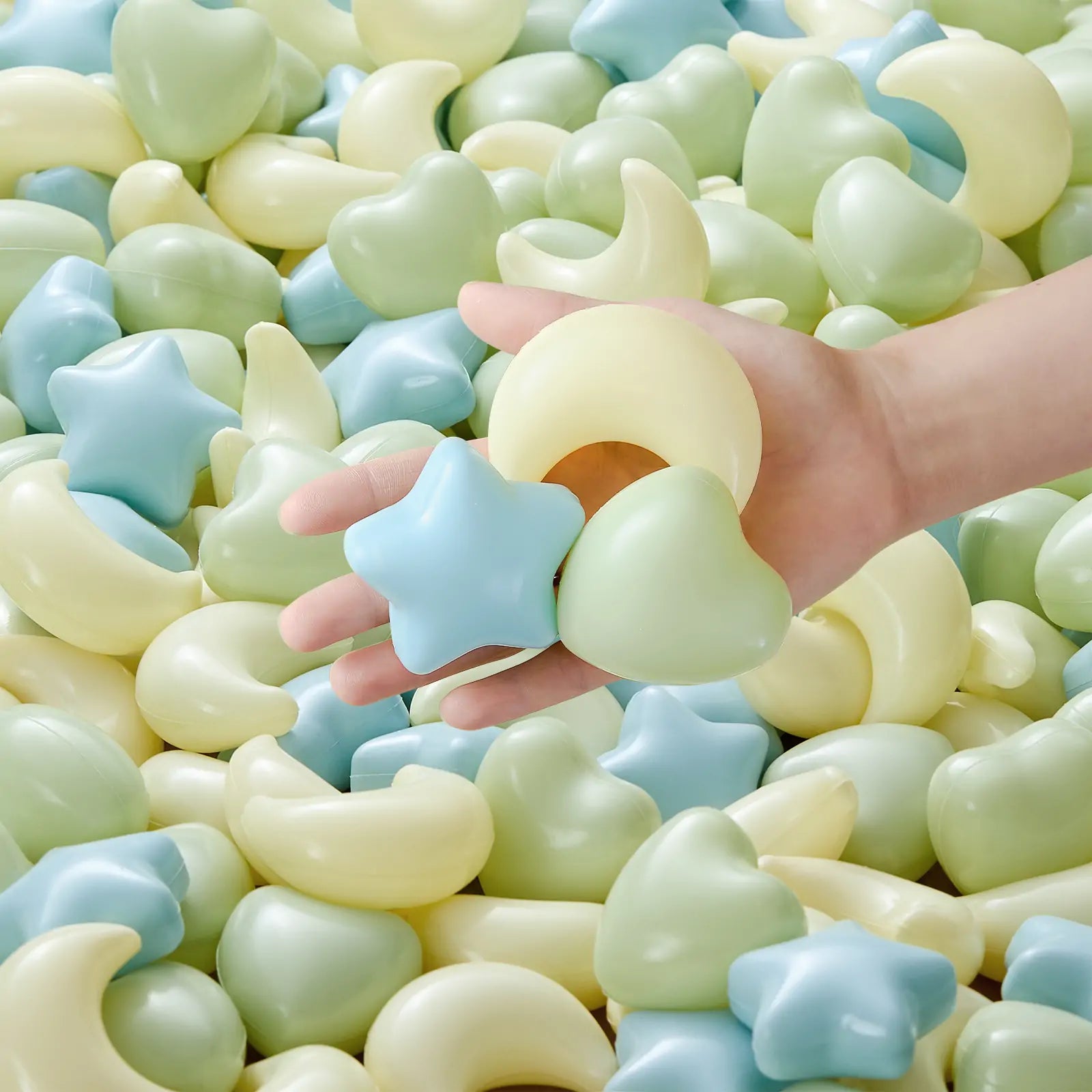 A hand holds pastel plastic shapes—a blue star, yellow moon, and green heart—from the Tiny Land® Green Adventure 3-in-1 Play Tunnel & Fun-Shapes Ocean Ball Set above soft, soothingly colored shapes for sensory play.