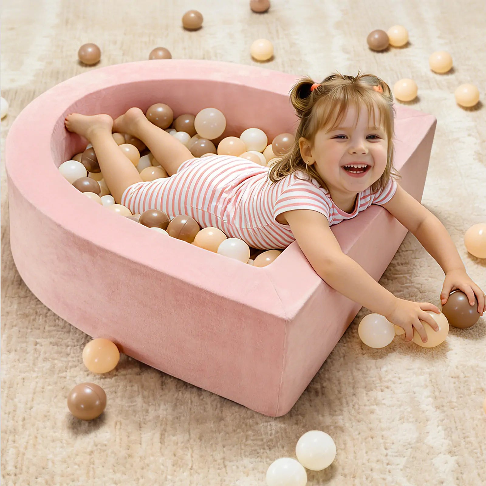 A smiling toddler in a striped outfit lies inside the Tiny Land® Violet PlayNest Climbing Ball Pit Set with Tunnel, surrounded by beige, brown, and white plastic balls on a light carpeted floor—creating an inviting indoor playground scene from Tiny Land.|Pink