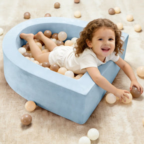 A young child smiles while lying in the Tiny Land® PlayNest Ball Pit with 200 beige, white, and brown ocean balls by Tiny Land, set on a light-colored carpet.