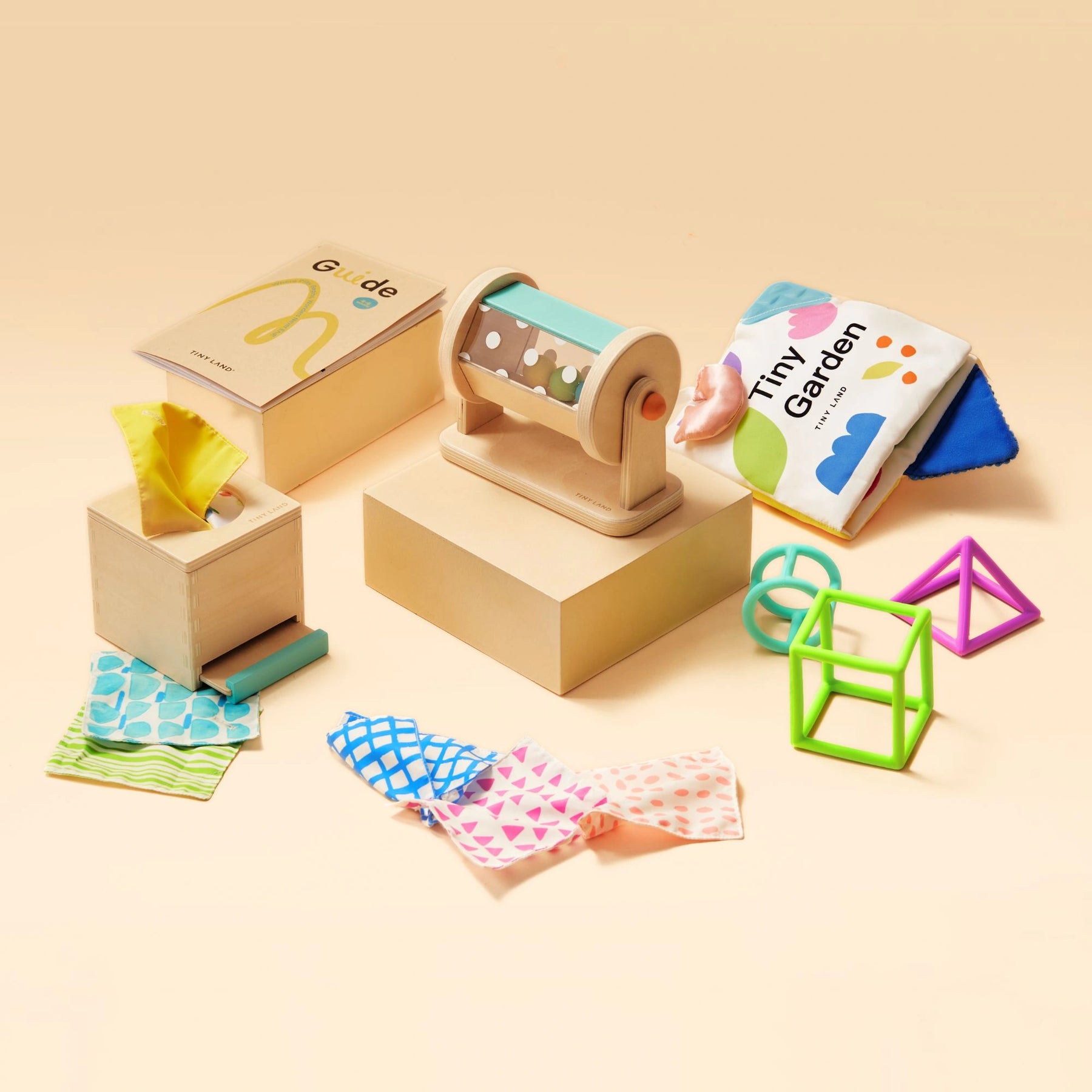 The Tiny Land Brain & Hands Playset — Montessori Box (4–6 Months) + Activity Cube features developmental toys like colorful fabric squares, textured blocks, soft books, interlocking shapes, and a wooden rolling toy arranged on a light beige surface.
