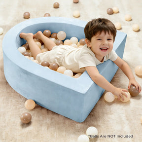 A young child smiles in a blue, heart-shaped Tiny Land® PlayNest Ball Pit (Ocean Ball Not Included) filled with beige, brown, and white plastic balls on a tan carpet. Some balls are on the floor. *Balls are NOT included.|blue
