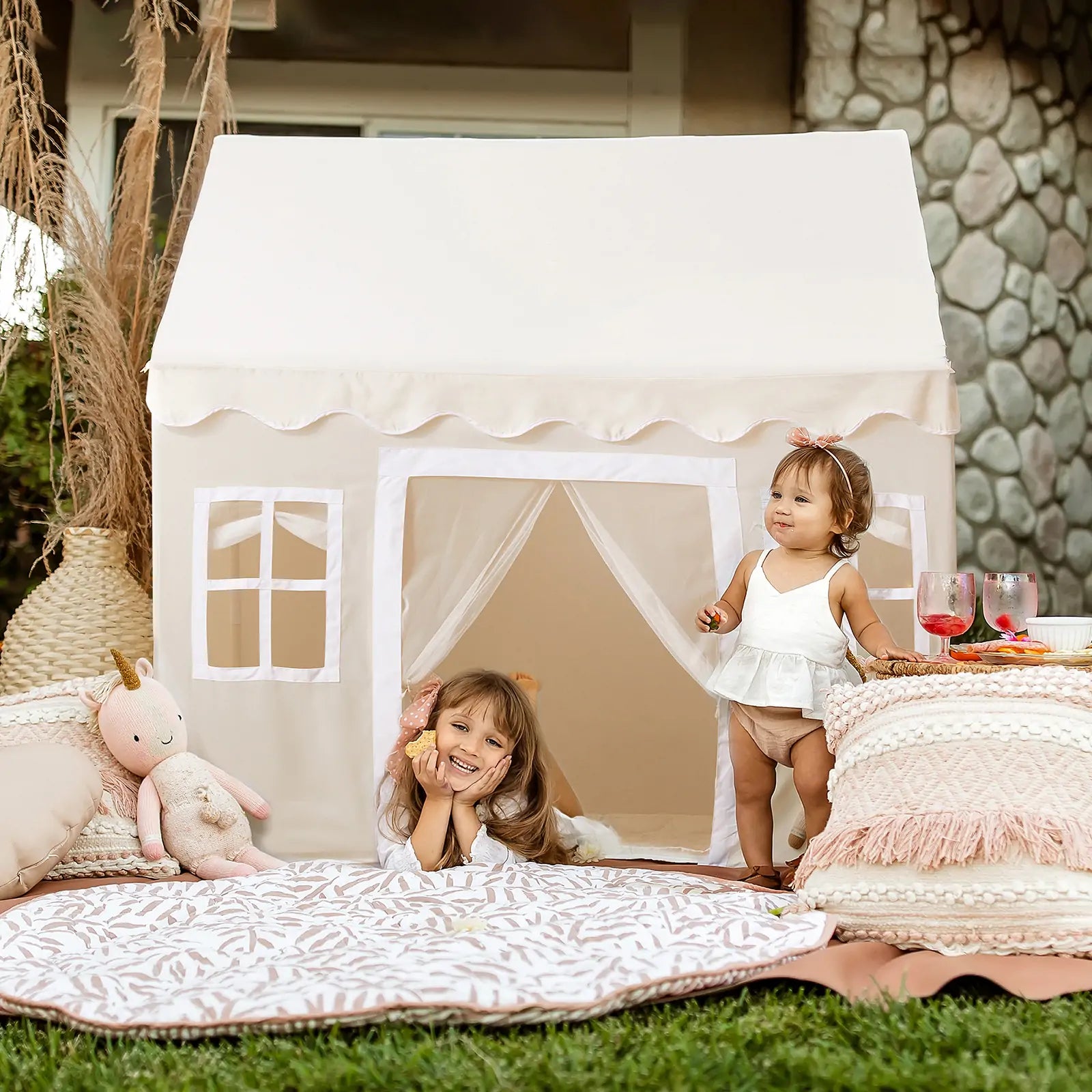 Two children enjoy the Tiny Land Bright Play & Art Set—Colorful Easel + Cream Playhouse with Lights. One girl smiles inside while the other stands at the entrance, surrounded by cushions, a plush toy, and drinks in this inviting indoor-outdoor setting|white