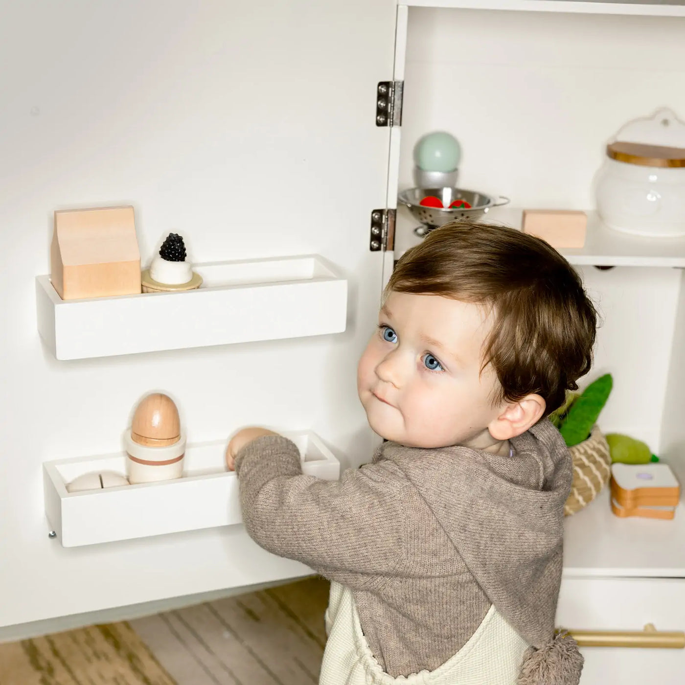 A toddler with blue eyes and brown hair in a gray hoodie plays with Tiny Land’s Food-Filled Kitchen Gift Set: Storage Kitchen & Fridge & Organic Food Collection, reaching into the toy fridge shelf of the kids kitchen set.