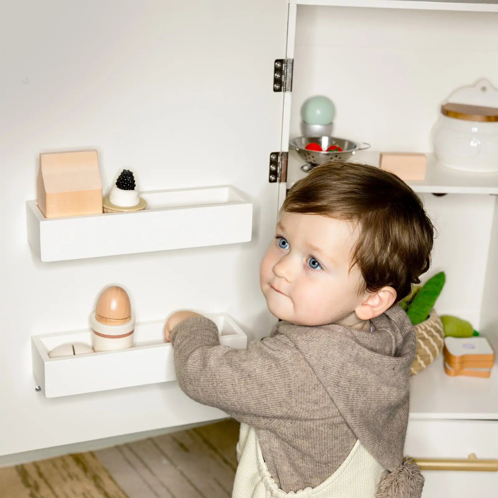 A toddler with blue eyes and brown hair in a gray hoodie plays with Tiny Land’s Food-Filled Kitchen Gift Set: Storage Kitchen & Fridge & Organic Food Collection, reaching into the toy fridge shelf of the kids kitchen set.